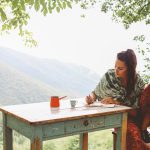 Vintage toned image of a young brunette woman relaxing in her weekend house. Sitting by the table, writing, on the wooden terrace in front yard, enjoying a beautiful view into the forest and the mountains.